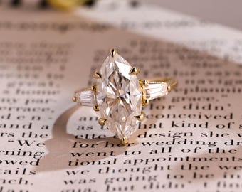May include: A gold engagement ring with a large, clear, marquise-cut diamond as the centerpiece. The ring has two smaller, rectangular diamonds on either side of the main stone. The ring is set on a textured, off-white surface.