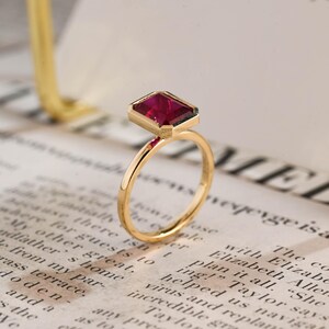 May include: A gold ring featuring a rectangular, deep pink gemstone set in a bezel setting. The band is thin and polished, showcasing a smooth, metallic finish that complements the vibrant, faceted stone held securely at the top of the ring.