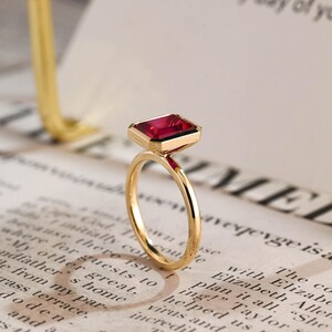 May include: A gold ring featuring a rectangular, emerald-cut deep red gemstone set in a polished gold bezel. The band is thin and circular, crafted from smooth, reflective gold metal, showcasing a clean, minimalist design with a vibrant red stone.