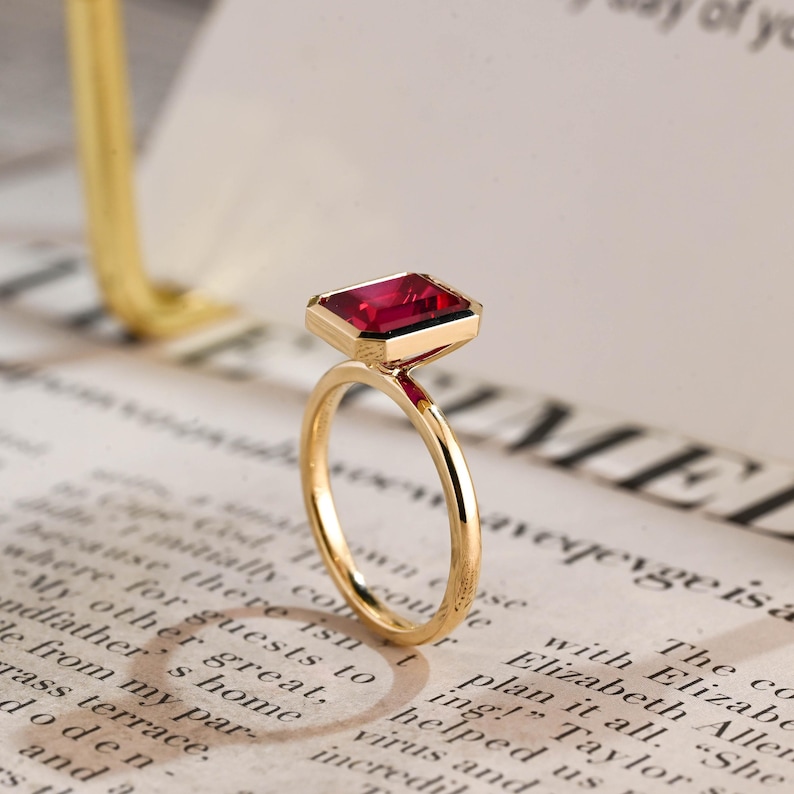 May include: A gold ring featuring a rectangular, emerald-cut deep red gemstone set in a polished gold bezel. The band is thin and circular, crafted from smooth, reflective gold metal, showcasing a clean, minimalist design with a vibrant red stone.