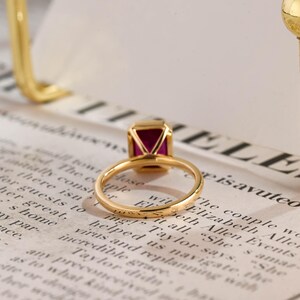 May include: A gold ring featuring a rectangular, deep pink gemstone set in a four-prong setting. The band is thin and polished, showcasing a smooth, metallic finish. The ring is positioned to highlight the vibrant colour and clarity of the stone.