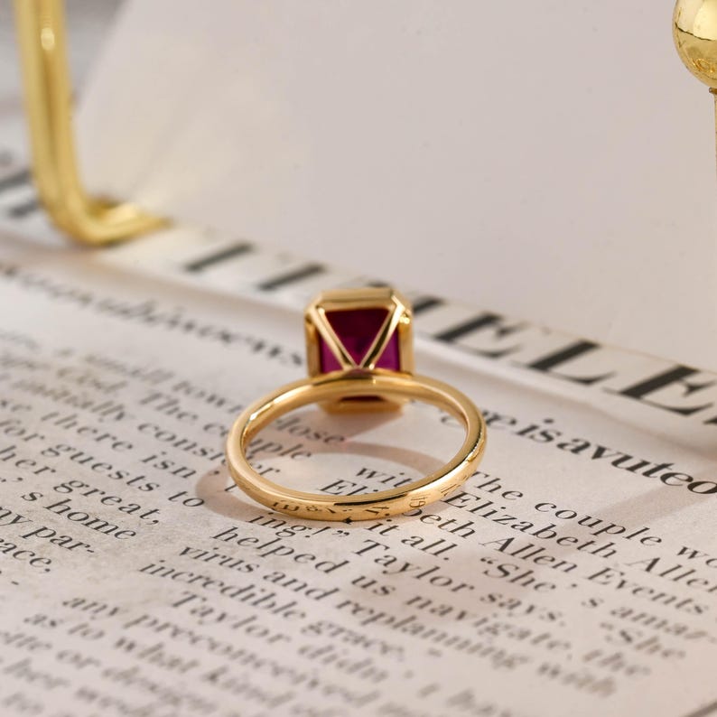May include: A gold ring featuring a rectangular, deep pink gemstone set in a four-prong setting. The band is thin and polished, showcasing a smooth, metallic finish. The ring is positioned to highlight the vibrant colour and clarity of the stone.
