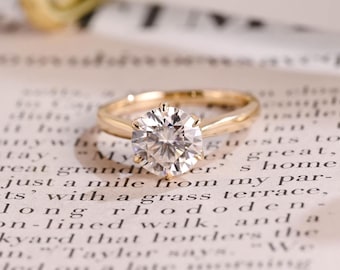 Puede incluir: Un anillo de oro con una piedra preciosa transparente de talla brillante redonda, sujeta por un engaste de seis garras. La banda es lisa y pulida, mostrando un diseño solitario clásico que descansa sobre una superficie con texto negro.