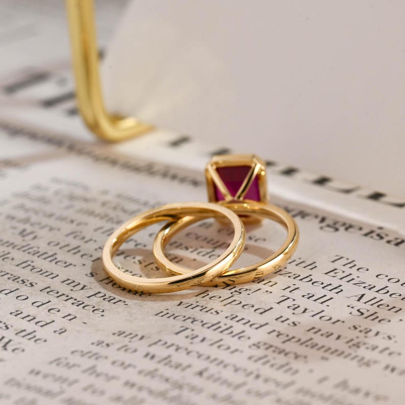 May include: A gold ring set consisting of a thin, plain gold band and a second gold band featuring a rectangular, deep pink gemstone set in a gold frame. The rings are positioned together, showcasing the polished metal and the vibrant stone.