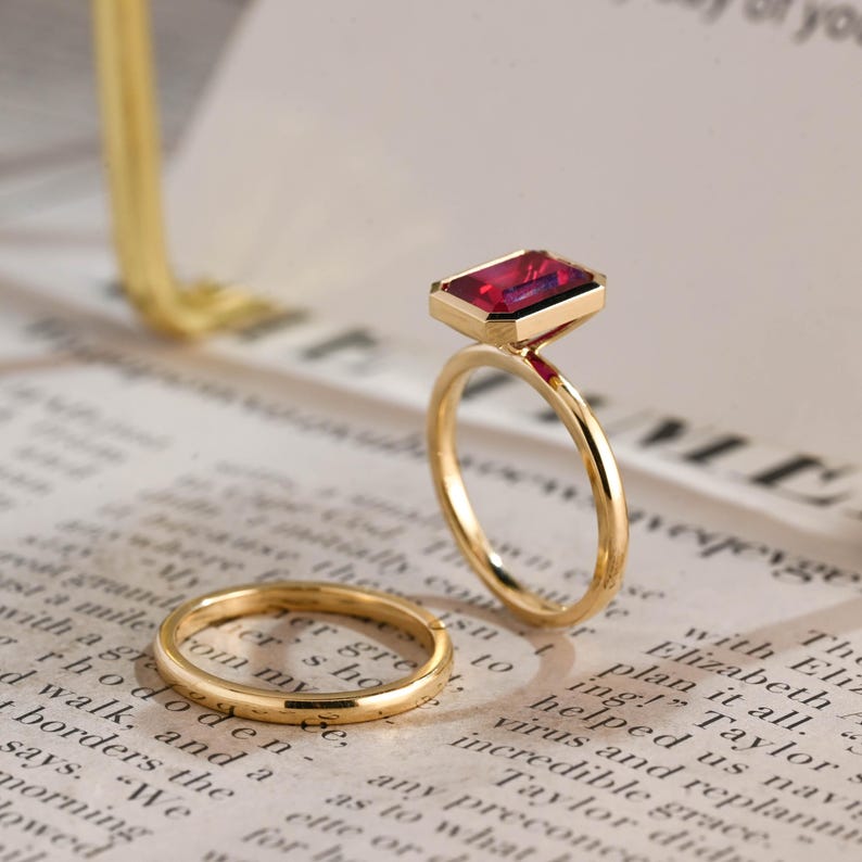 May include: A gold ring set consisting of one plain thin band and one ring featuring a rectangular emerald-cut deep red gemstone set in a gold bezel. The rings are positioned upright and flat on a surface with printed text.