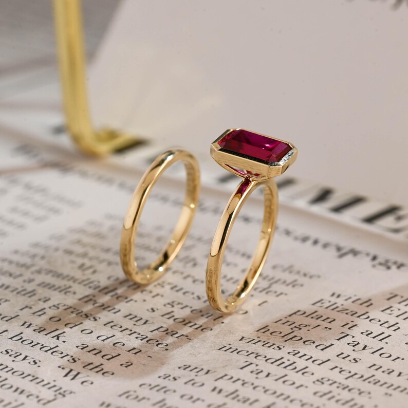 May include: A gold ring set consisting of a plain thin band and a second band featuring a rectangular, emerald-cut, deep pink gemstone set in a gold bezel. The rings are positioned upright, showcasing the polished metal finish and the vibrant stone.