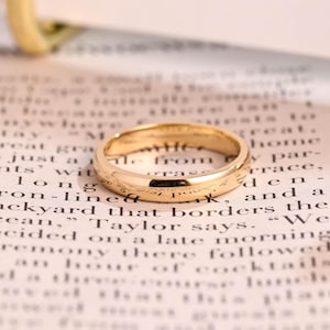 May include: A gold wedding ring with a smooth, polished surface. The ring is placed on a page of printed text, offering a classic and refined look. The ring's design is simple and timeless.