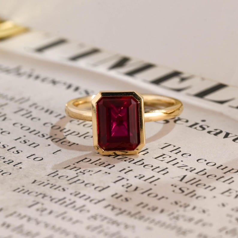 May include: A gold ring featuring a rectangular, emerald-cut deep red gemstone set in a simple polished gold band. The ring is positioned on a page of printed text, highlighting the vibrant colour and faceted surface of the central stone.