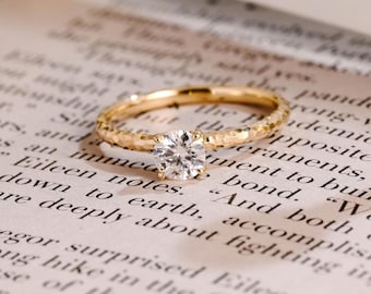 Può includere: Un anello in oro con una singola pietra preziosa trasparente a taglio brillante rotondo, fissata con quattro griffe. La fascia dell'anello presenta una finitura martellata e testurizzata che cattura la luce, creando una superficie riflettente.
