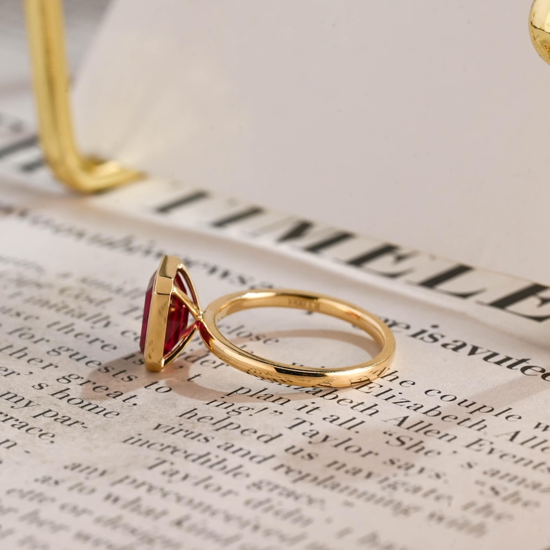 May include: A gold ring featuring a rectangular, deep red gemstone set in a four-prong setting. The band is thin and polished, showcasing a smooth, rounded profile. The ring is positioned at an angle, highlighting the vibrant red stone and the metallic lustre of the gold band against a printed background.
