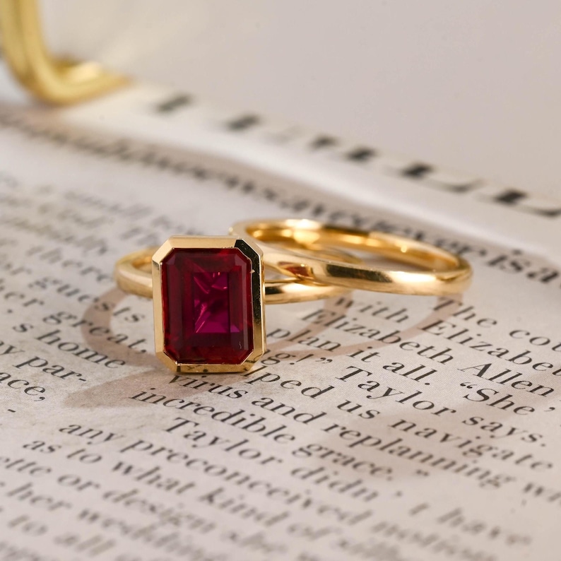 May include: A gold ring featuring a rectangular, emerald-cut deep red gemstone set in a bezel setting. The band is thin, polished, and circular, showcasing a smooth, metallic finish that reflects light across its surface and the vibrant red stone.