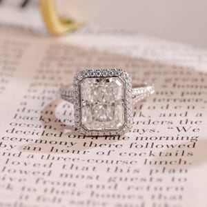 May include: A silver engagement ring featuring a large, rectangular-cut diamond surrounded by a halo of smaller stones. The ring is set on a printed page, showcasing its intricate design and sparkle.