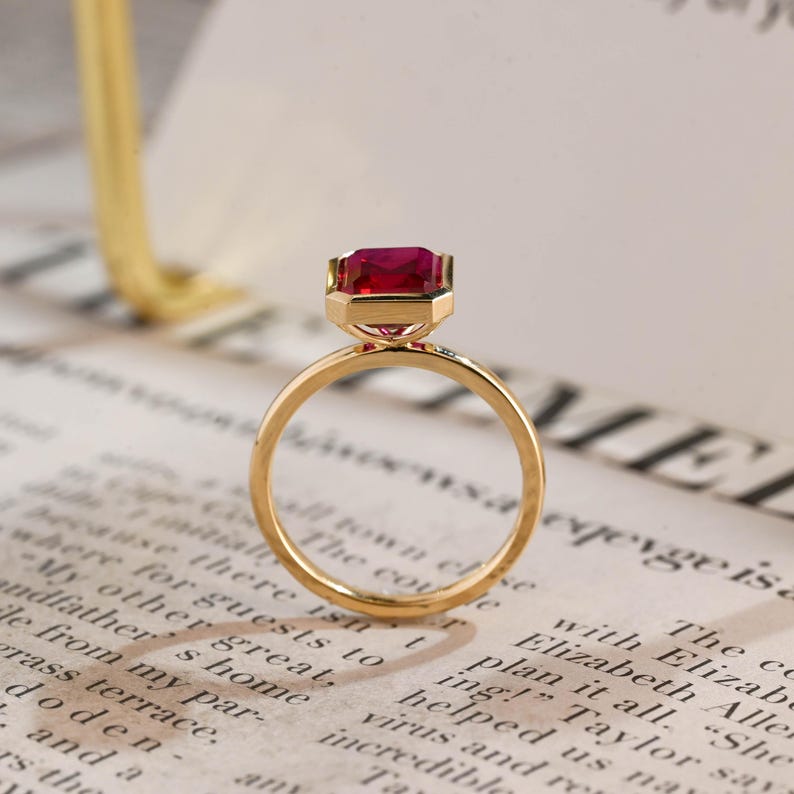 May include: A gold ring featuring a rectangular, faceted, deep red gemstone set in a polished gold bezel. The ring has a thin, smooth, circular gold band. The design highlights the vibrant contrast between the metallic gold and the red stone.