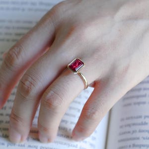 May include: A gold ring featuring a rectangular, emerald-cut red gemstone set in a bezel setting. The ring has a thin, polished gold band that encircles the finger, showcasing the vibrant red stone as the central focal point of the jewellery piece.