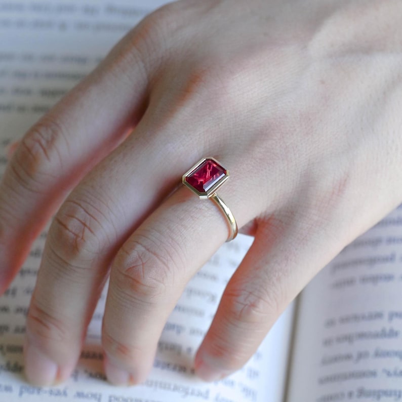 May include: A gold ring featuring a rectangular, emerald-cut red gemstone set in a bezel setting. The ring has a thin, polished gold band that encircles the finger, showcasing the vibrant red stone as the central focal point of the jewellery piece.