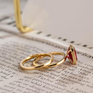 May include: A gold ring set consisting of a thin, plain band and a second band featuring a prominent, rectangular, deep red gemstone set in a gold frame. The rings are positioned together, showcasing the polished gold metal and the vibrant red stone.