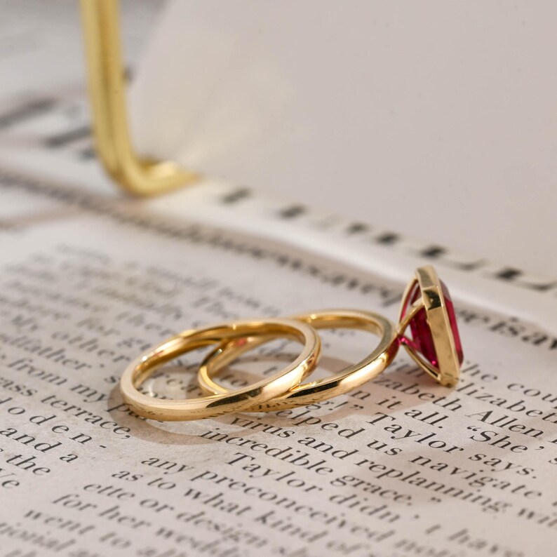 May include: A gold ring set consisting of a thin, plain band and a second band featuring a prominent, rectangular, deep red gemstone set in a gold frame. The rings are positioned together, showcasing the polished gold metal and the vibrant red stone.