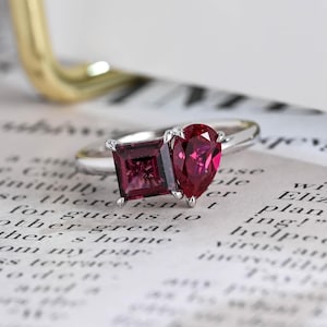 Pode incluir: Um anel de prata com duas pedras preciosas vermelhas: uma de corte quadrado e outra em forma de pêra. O anel está sobre um fundo de texto impresso, com um objeto dourado ao fundo.