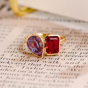 May include: A gold ring featuring two distinct gemstones: a pear-shaped purple stone and a rectangular-cut red stone. The stones are set side-by-side in a polished gold band, creating a dual-stone design that highlights the contrasting colours.