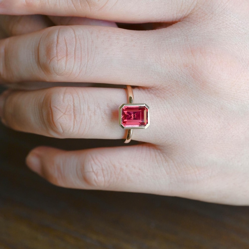 May include: A gold ring featuring a rectangular, emerald-cut vibrant pink gemstone set in a smooth bezel setting. The band is thin and polished, showcasing the faceted stone which sits horizontally across the finger, highlighting the clear pink hue.