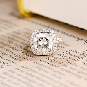 Op de afbeelding: Een zilverkleurige ring met een grote, kussengeslepen centrale steen omringd door een halo van kleine, ronde briljantgeslepen stenen. De ringband is gespleten en versierd met extra kleine, ronde briljantgeslepen stenen aan de zijkanten.
