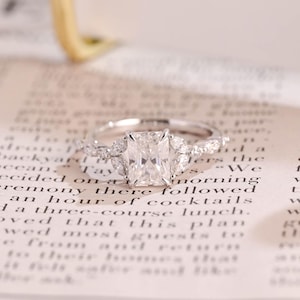 Puede incluir: Un anillo de compromiso plateado con una gran piedra preciosa rectangular y transparente. El anillo tiene piedras de acento más pequeñas y claras en la banda y alrededor de la piedra principal. Descansa sobre una página de texto impreso.