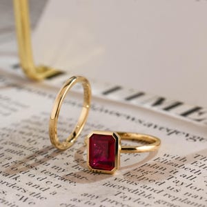 May include: A gold ring set consisting of a plain, thin gold band and a second gold band featuring a rectangular, emerald-cut red gemstone set in a bezel setting. The rings are polished and have a smooth, reflective metallic finish.