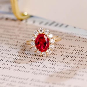 Puede incluir: Un anillo de oro con una piedra preciosa roja central de corte ovalado, rodeada por un halo de pequeñas piedras blancas redondas y transparentes. La banda es fina y de oro pulido, con un diseño clásico y una piedra roja vibrante.