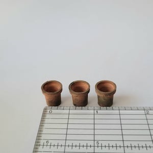 May include: Three small terracotta pots, each with a slightly different shade of brown, are lined up on a white surface with a ruler behind them.
