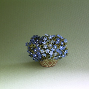 以下が含まれることがあります： 青い星型の花と緑の葉を持つ、小さな鉢植えの植物。 鉢は薄茶色で、ひし形の模様があります。 背景は無地のオリーブグリーンです。