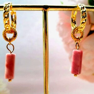 Può includere: Un paio di orecchini a cerchio color oro con un design a catena. Ogni orecchino presenta un piccolo pendente rettangolare rosa. Gli orecchini sono esposti su un supporto dorato, con un morbido sfondo floreale.