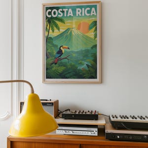 Può includere: Un poster incorniciato per il Costa Rica, raffigurante un tucano su un ramo con un vulcano e un tramonto. Il poster è in una cornice di legno e appeso a una parete bianca. Il testo "COSTA RICA" è in alto.