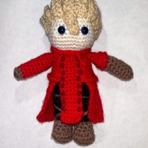 May include: A handmade crochet doll with a light tan head and hair, blue eyes, and a red coat. The doll's arms and legs are brown, and it has a white face. The red coat has several buttons and a detailed design. The doll is standing upright.