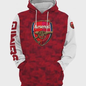 Może przedstawiać: Czerwona i biała bluza z kapturem Arsenal z herbem drużyny z przodu. Herb przedstawia armatę i napis "Arsenal". Rękawy są białe, z napisem "Gunners" wydrukowanym pionowo na lewym rękawie. Bluza ma kaptur ze sznurkiem i przednią kieszeń.