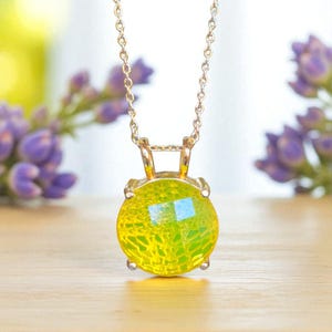 May include: A gold-toned necklace with a round, yellow-green pendant. The pendant has a textured, faceted appearance, and the chain is delicate. The background features blurred purple flowers and a wooden surface.