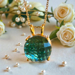 May include: A gold-toned necklace featuring a round, faceted pendant with shades of teal and blue. The pendant is suspended from a delicate chain. The background includes white roses, pearls, and greenery, creating an elegant aesthetic.