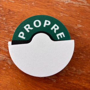 May include: A white and green sign with the word "PROPRE" in white letters. The sign is a half-circle shape with a cutout at the bottom. The sign is on a wooden surface.