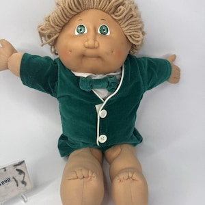 Pode incluir: Uma boneca Cabbage Patch Kid vintage com pele bronzeada, olhos verdes e cabelo loiro de fio. A boneca está vestindo uma jaqueta de veludo verde escuro com uma camisa branca e uma gravata borboleta. As pernas e os braços da boneca são feitos de tecido macio.
