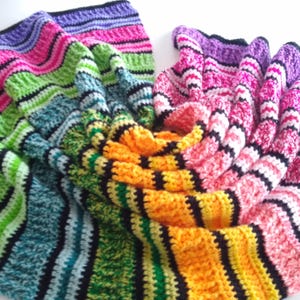 May include: A colorful, handmade crochet blanket with a striped pattern. The blanket features a variety of colors, including pink, green, blue, yellow, and purple, with black stripes. The blanket is folded and draped, showcasing its texture and design.