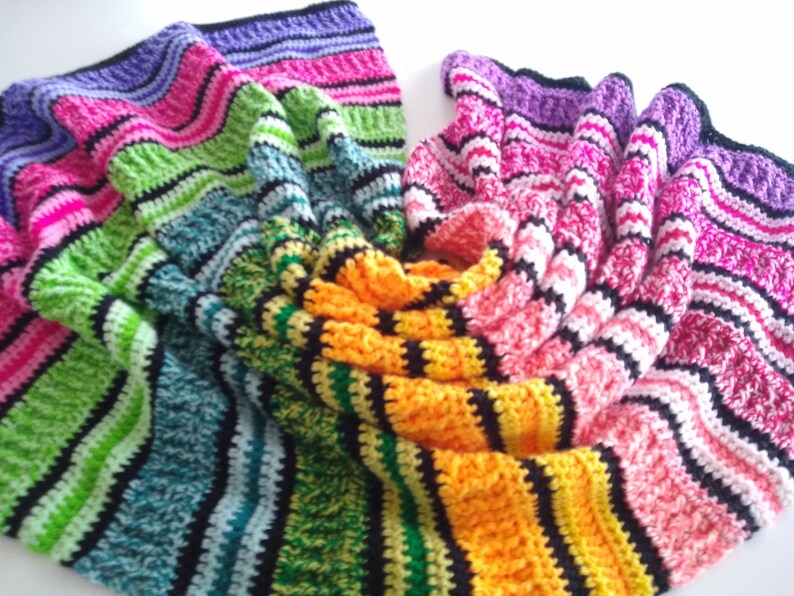 May include: A colorful, handmade crochet blanket with a striped pattern. The blanket features a variety of colors, including pink, green, blue, yellow, and purple, with black stripes. The blanket is folded and draped, showcasing its texture and design.