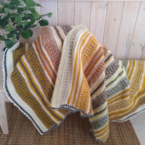 May include: A handmade crochet blanket with a striped pattern in shades of yellow, orange, brown, and cream. The blanket is draped over a wooden bench, showcasing its textured design and cozy appeal. The edges are finished with a white and navy blue border.