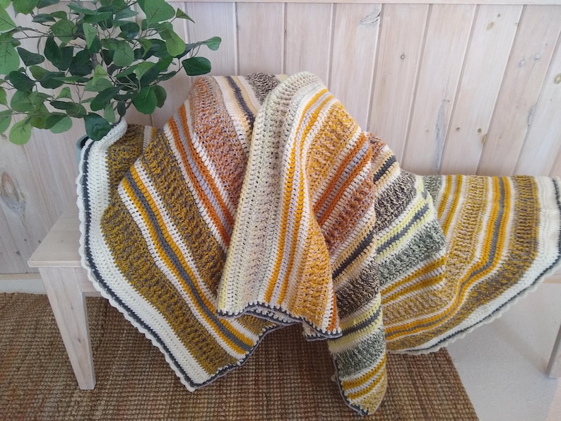 May include: A handmade crochet blanket with a striped pattern in shades of yellow, orange, brown, and cream. The blanket is draped over a wooden bench, showcasing its textured design and cozy appeal. The edges are finished with a white and navy blue border.