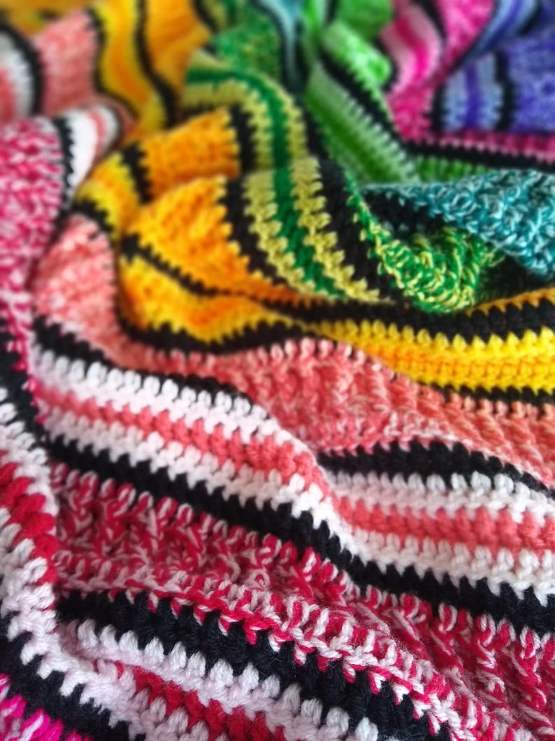 May include: A colorful, handmade crochet blanket. The blanket features a vibrant mix of colors including red, pink, yellow, green, and blue. The crochet pattern includes black stripes that separate the different color sections.