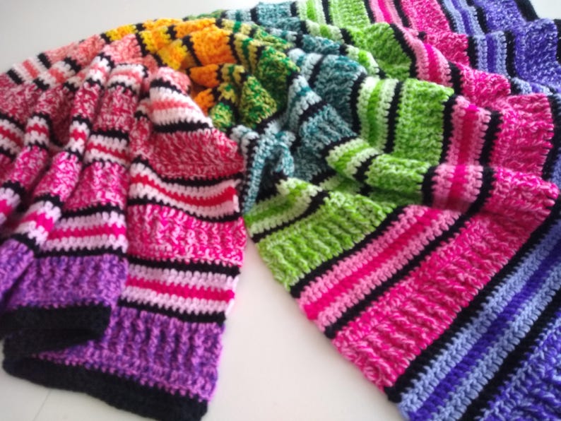 May include: A colorful, handmade crochet blanket with a striped pattern. The blanket features a vibrant array of colors, including pink, purple, green, yellow, and blue, with black stripes separating the color blocks. The blanket is folded and draped.