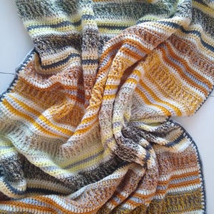 May include: A handmade crochet blanket with a striped pattern in shades of yellow, brown, gray, and white. The blanket is folded and draped, showcasing the texture and color variations. The edges are finished with a dark blue border.