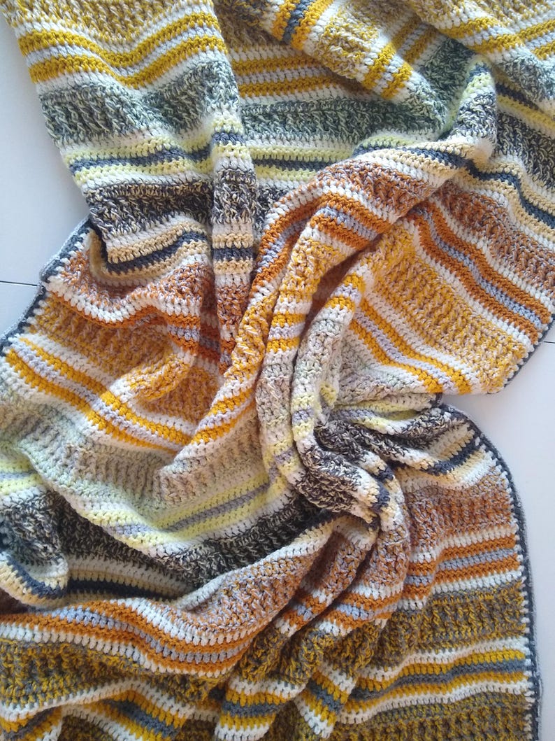 May include: A handmade crochet blanket with a striped pattern in shades of yellow, brown, gray, and white. The blanket is folded and draped, showcasing the texture and color variations. The edges are finished with a dark blue border.