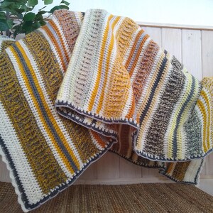 May include: A handmade crochet blanket with a striped pattern in shades of cream, yellow, gray, and brown. The blanket has a scalloped edge and is draped over a wooden bench. The blanket is approximately 60 inches wide and 70 inches long.