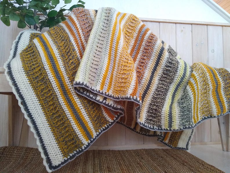 May include: A handmade crochet blanket with a striped pattern in shades of cream, yellow, gray, and brown. The blanket has a scalloped edge and is draped over a wooden bench. The blanket is approximately 60 inches wide and 70 inches long.