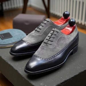 May include: Two-tone dress shoes with grey suede uppers and black leather toe caps and heels. The shoes feature black laces and decorative perforations. Shoe trees are inserted. The shoes are displayed on a dark surface.