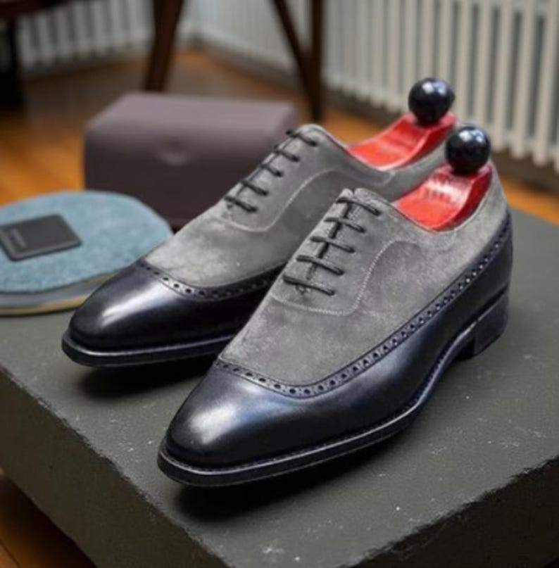 May include: Two-tone dress shoes with grey suede uppers and black leather toe caps and heels. The shoes feature black laces and decorative perforations. Shoe trees are inserted. The shoes are displayed on a dark surface.